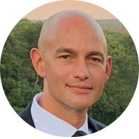 A bald man in a suit and tie is smiling outdoors with trees in the background, perhaps enjoying a refreshing break from his hand therapy session in Nesconset.